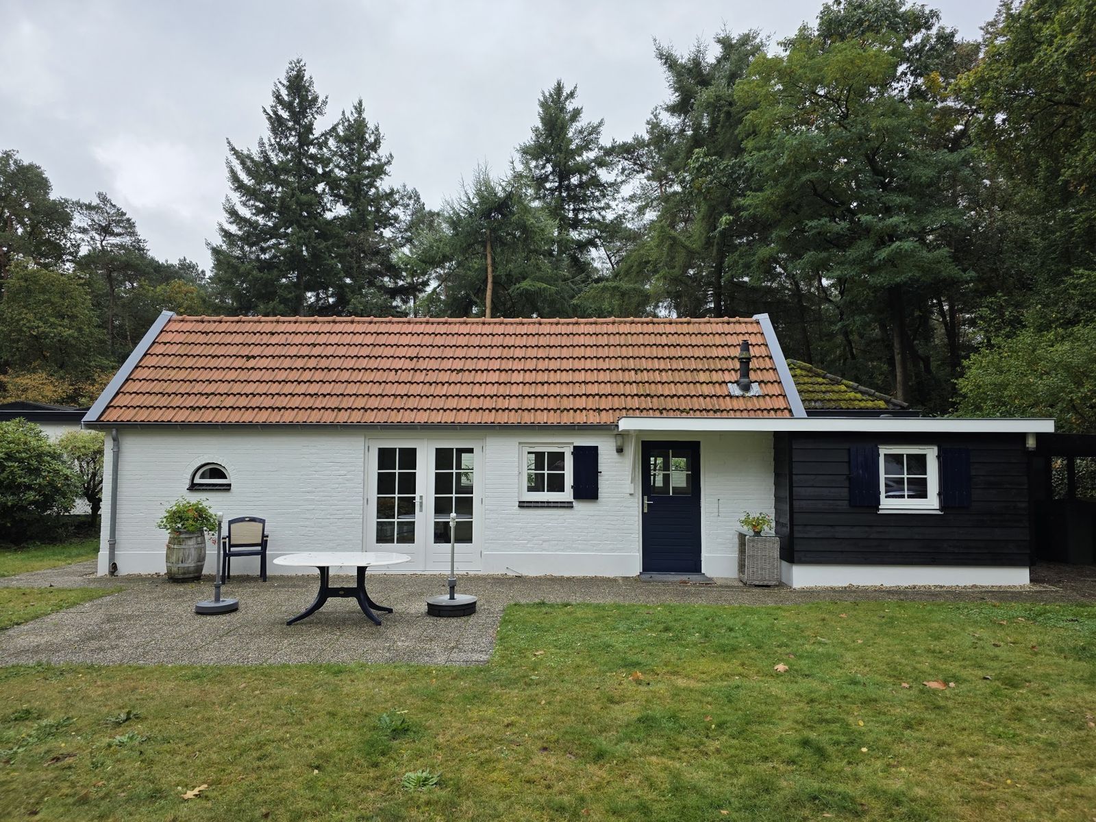 Wit geschilderd huisje in een bosrijke tuin, met zwarte deur, zwarte luiken en een zwarte houten aanbouw