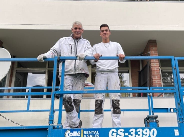 Willy en Alex van der Beek in witte schildersoveralls op een hoogwerker voor een gebouw