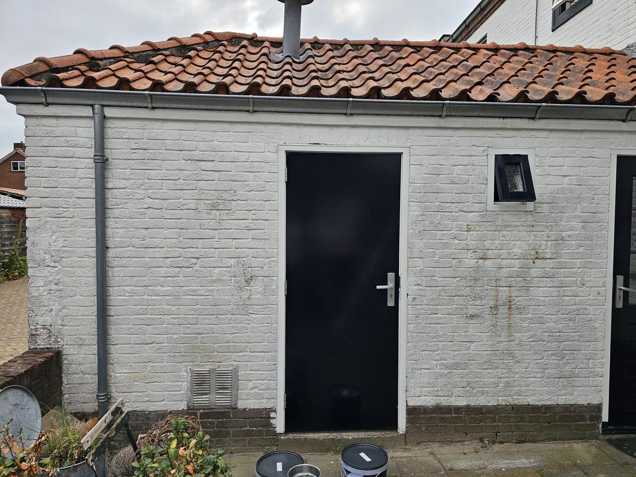 Voor: wit geschilderd schuurtje met verweerde, vlekkerige muren en roestsporen, verfblikken ervoor